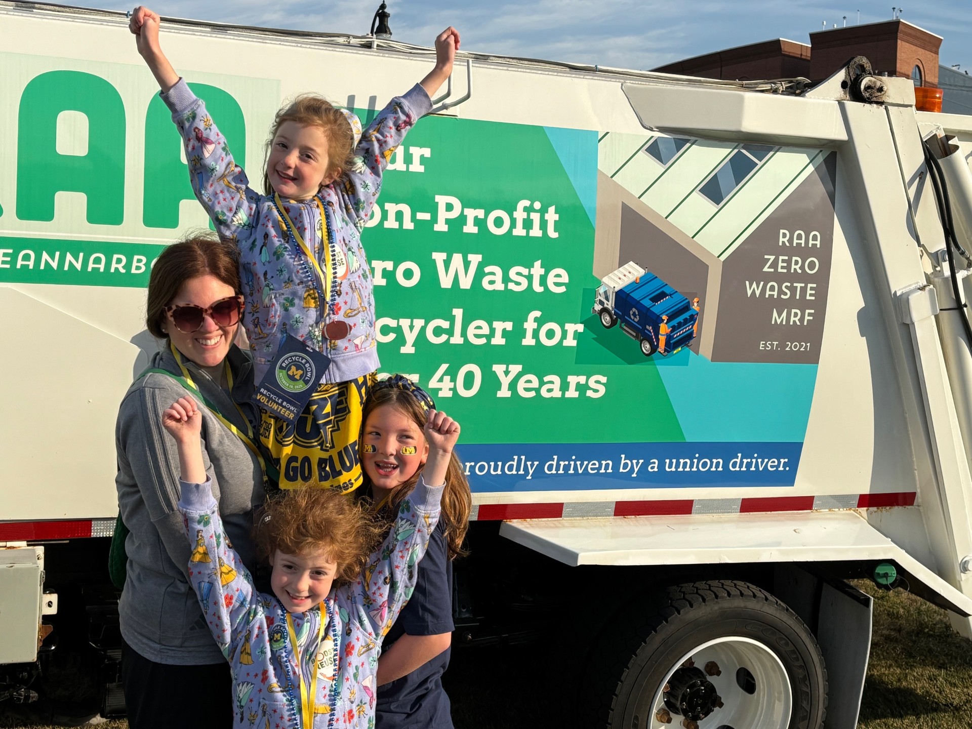 A family celebrates in front of a recycling truck during the University of Michigan Recycle Bowl, highlighting PET bottle recycling and game-day sustainability efforts.