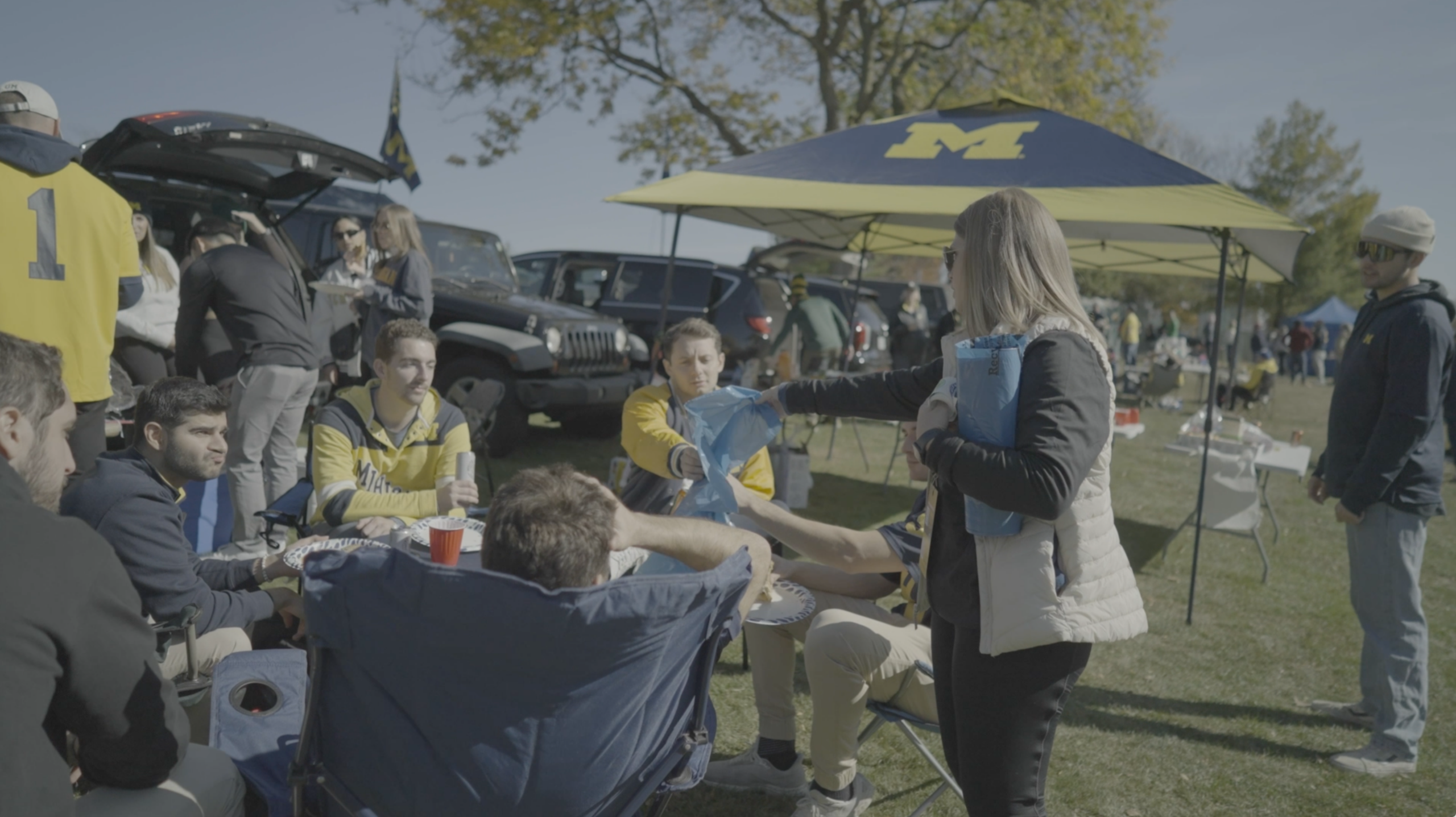 A Recycle Bowl volunteer hands blue recycling bags to University of Michigan football fans at a tailgate, encouraging PET bottle recycling before the game.