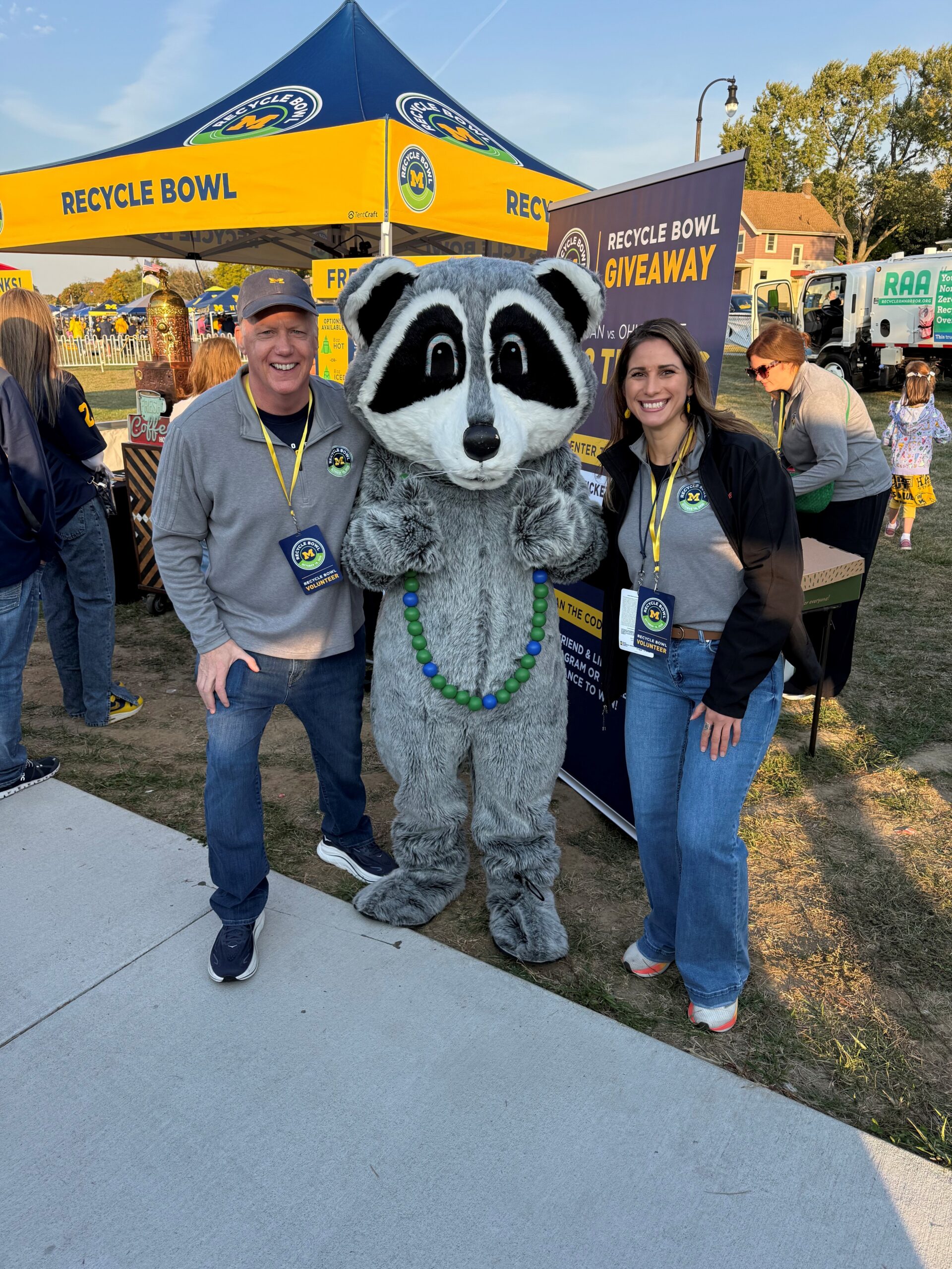 Two Recycle Bowl volunteers pose with the Recycling Raccoon mascot at a University of Michigan football tailgate.